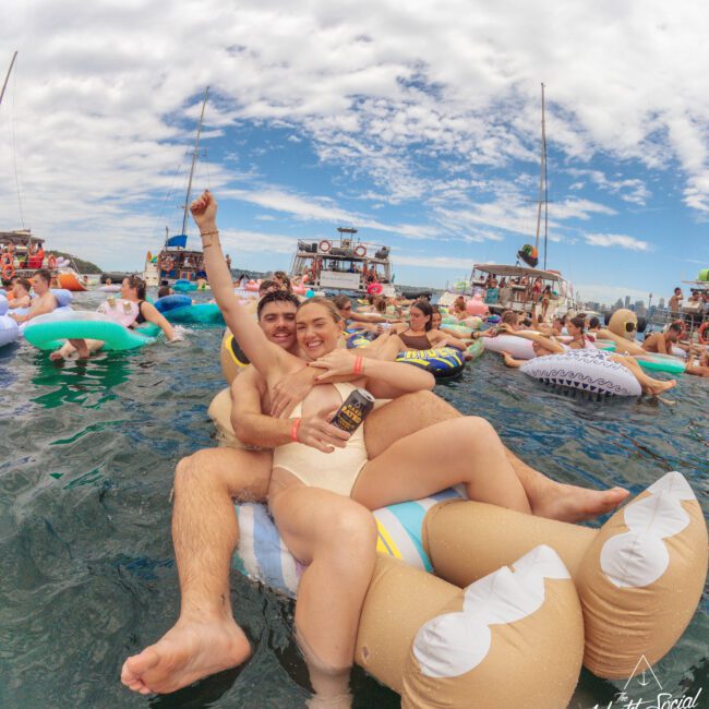 A couple in swimsuits smiles and poses on a large inflatable float in the water, surrounded by other people on colorful inflatables at a lively outdoor pool party. Boats and a partly cloudy sky are in the background.