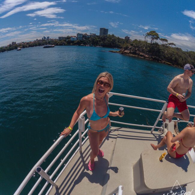 A woman in a blue bikini smiles and poses with a drink on a boat deck, with water and city buildings in the background. Two other people sit nearby under a sunny, blue sky.