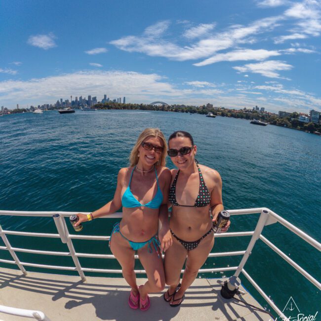 Two women in bikinis smiling and holding drinks stand on the deck of a boat with blue water and a city skyline in the background under a sunny sky.