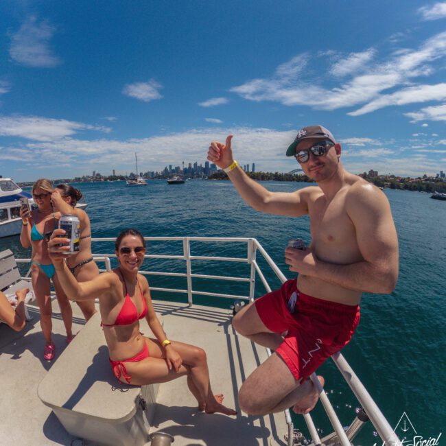 Two people in swimsuits smile and pose on a boat, holding drinks with the ocean and city skyline in the background. Other people relax nearby under a sunny blue sky.