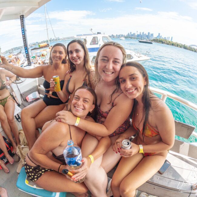 A group of five women in swimsuits smile and pose together on a boat with drinks in hand. Other people and boats are visible in the background on the water with a city skyline in the distance.