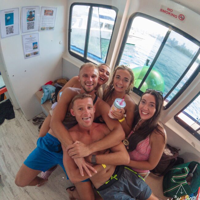 Five smiling young adults in swimsuits pose closely together inside a boat, with water and another boat visible through the window. One person holds a drink. The atmosphere is casual and joyful.