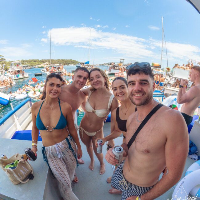 Five young adults in swimsuits smile for a group photo on a boat during a lively yacht party, with more people, boats, and blue skies visible in the background. Some are holding drinks, and the mood is festive and fun.
