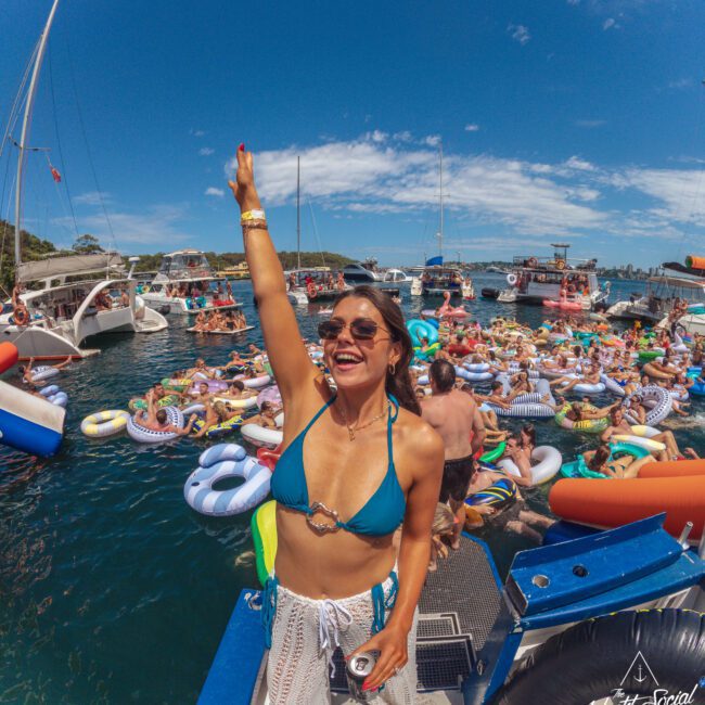 A woman in a blue bikini top and sunglasses smiles and raises her arm while standing on a boat, with a festive crowd of people on colorful floats and boats in the water behind her under a sunny sky.