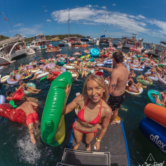 A woman in a red bikini with a green inflatable steps onto a floating dock, smiling at the camera. Behind her, many people relax on colorful inflatables on the water, surrounded by boats on a sunny day.
