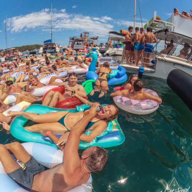 A large group of people enjoy a party in the water, lounging on colorful inflatable floats near anchored boats under a sunny sky. Several people are laughing and having drinks, creating a lively, festive atmosphere.