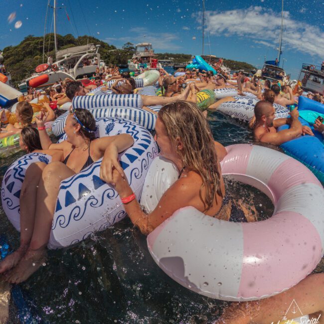 A large group of people float on colorful inflatable rings in the sea, surrounded by boats under a sunny blue sky. Water splashes in the air as everyone enjoys the lively outdoor scene.
