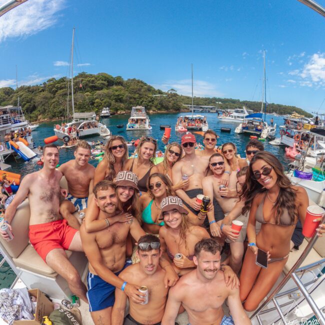 A group of people in swimsuits smile and pose together on a boat, holding drinks, with several other boats and green trees visible in the background on a sunny day.