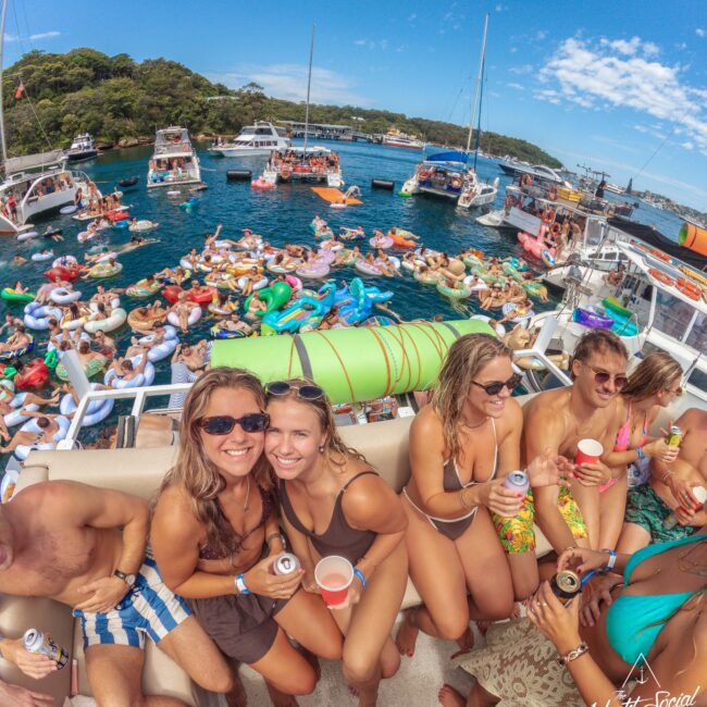 A large group of people enjoying a lively boat party; some are on a crowded deck smiling and holding drinks, while many others relax on colorful inflatables in the blue water surrounded by more boats.