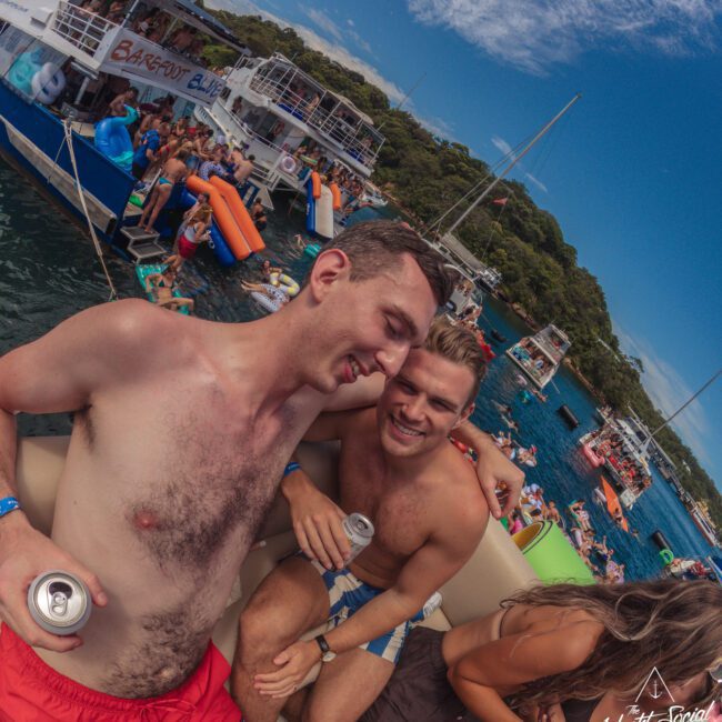 Two young men smile and pose with drinks at a lively boat party. Other people swim, lounge on inflatables, and gather on nearby boats under a bright blue sky. The atmosphere is festive and fun.
