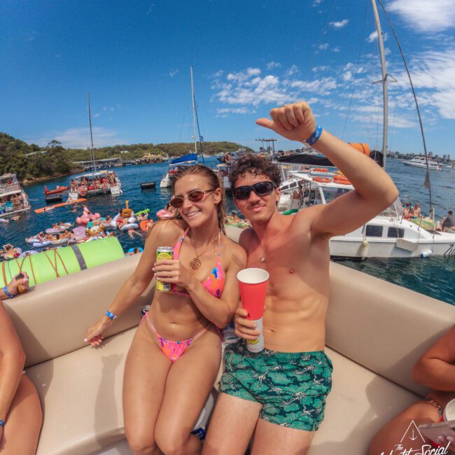 A woman in a pink bikini and a man in green patterned shorts and sunglasses smile and pose on a yacht with drinks in hand. Other people, boats, and inflatables fill the sunny background on the water.