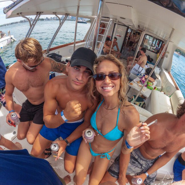 A group of young adults in swimwear smile and pose on a boat, holding drinks and enjoying a sunny day on the water. The sea and other boats are visible in the background.