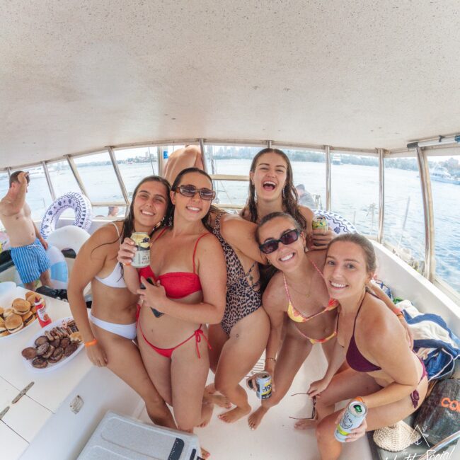 Five young women in swimsuits smile and pose with drinks on a boat, surrounded by snacks and the ocean visible through the windows. It's a sunny, cheerful atmosphere, suggesting a fun day out on the water.