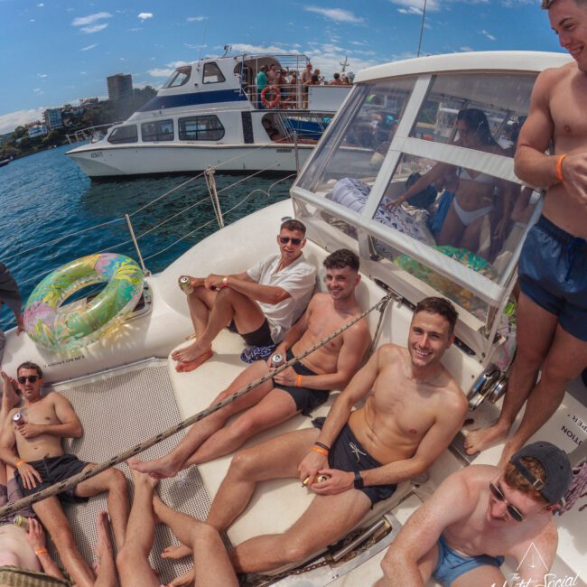 A group of young men in swimwear relax and smile on the deck of a boat in sunny weather, with another boat and blue water visible in the background. Some are seated while others recline, enjoying a casual outing.