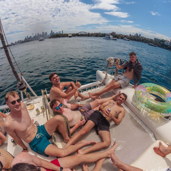 A group of young men relax shirtless on the netting of a catamaran, smiling and holding drinks. The boat floats on a harbor with a city skyline and blue sky with clouds in the background.