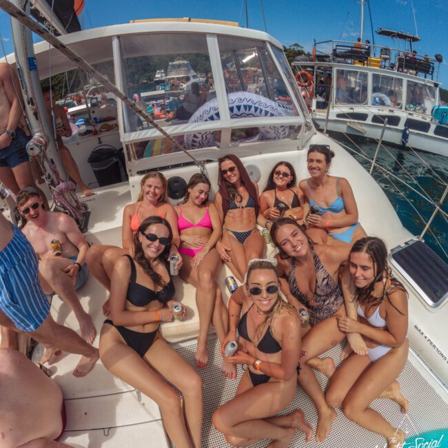 A group of smiling people in swimsuits sit and pose together on the deck of a boat, enjoying drinks under a sunny sky, with other boats and water visible in the background.