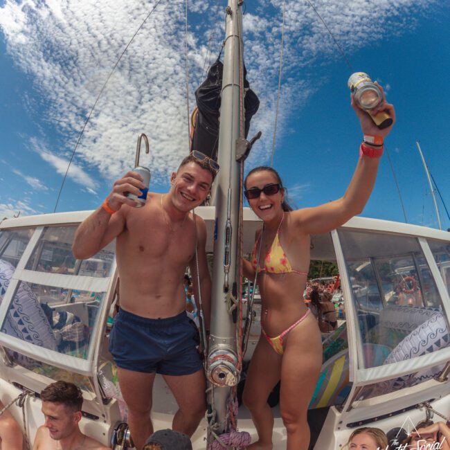Two people in swimsuits stand and smile on a boat, holding up cans and drinks. Other people sit and relax around them under a partly cloudy sky, enjoying a lively, sunny day on the water.