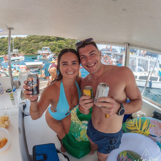 A smiling man and woman in swimwear hold drinks and pose for a photo on a boat, with people, other boats, and floaties visible in the water in the background. The mood is festive and summery.