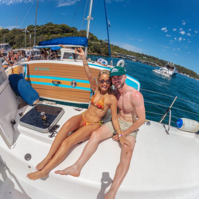 A smiling woman in a bikini and a man in swim trunks and a cap sit on the deck of a boat, with blue water and other boats in the background, enjoying a sunny day.
