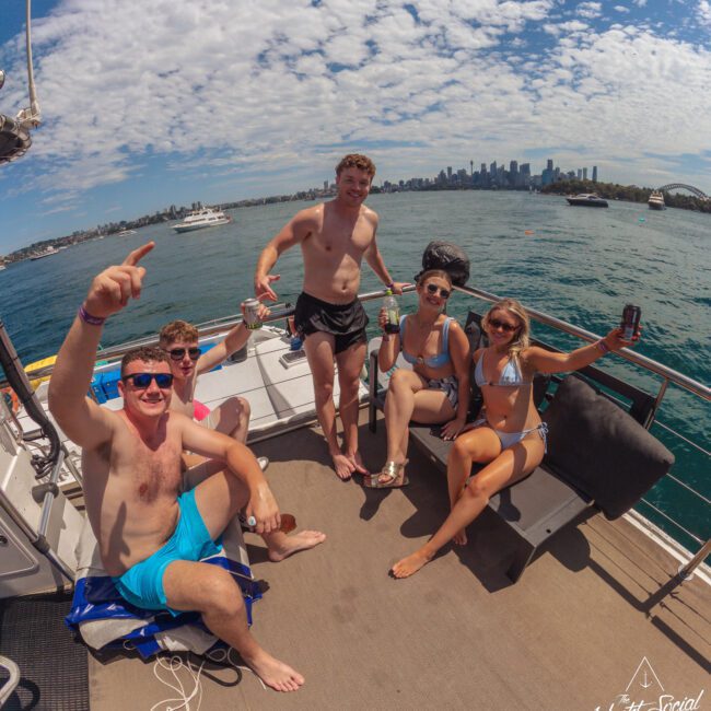 Five people in swimsuits sit and stand on a boat, smiling and holding drinks, with a city skyline and blue sky with clouds in the background. The atmosphere is relaxed and summery.