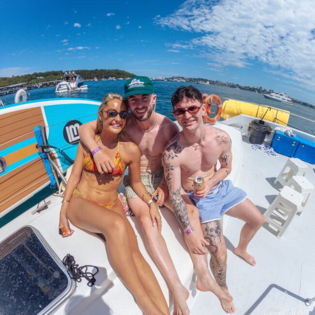Three smiling people in swimwear sit closely together on a boat under a sunny blue sky, with water, an inflatable structure, and other boats visible in the background.