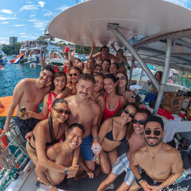 A large group of smiling people in swimwear pose for a selfie on a boat under sunny skies. The water and other boats are visible in the background, creating a fun, festive atmosphere.