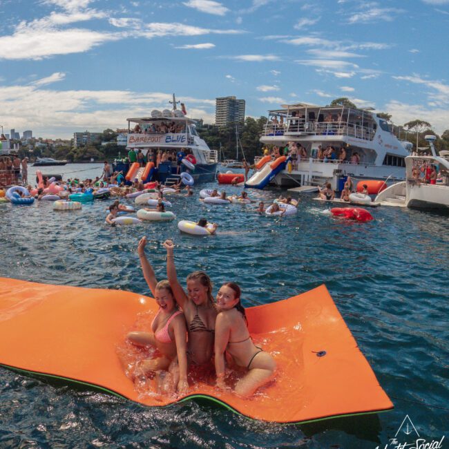 Three women in swimsuits pose on an orange floating mat in the water, with yachts and people on inflatables celebrating in the background under a sunny sky.