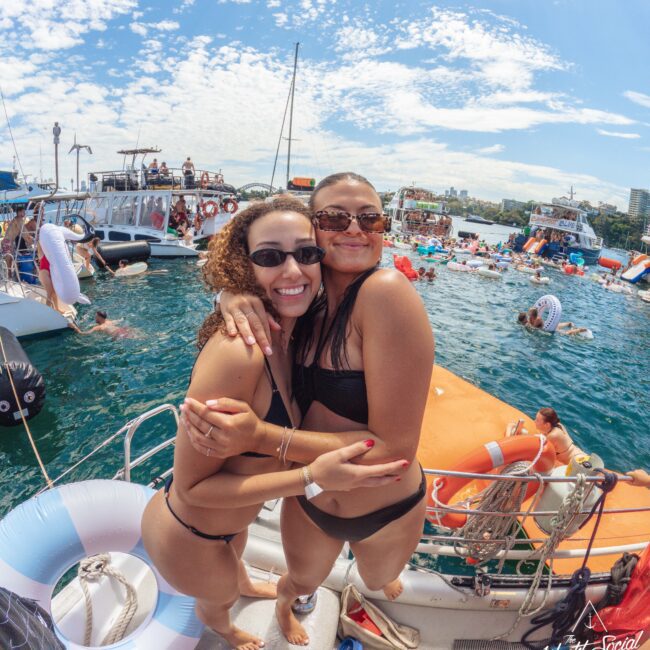 Two women in swimsuits smile and hug on the deck of a boat at a lively pool party, surrounded by people, floats, and other boats on a sunny day with blue skies and scattered clouds.