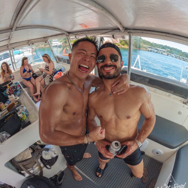 Two shirtless men smile and pose together on a boat; one holds a can and wears sunglasses. Other people in swimsuits relax nearby. The boat is on the water, with the shoreline visible in the background.