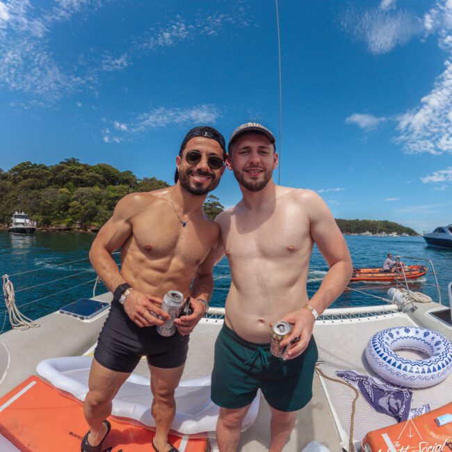 Two men in swim trunks stand on a boat, smiling and holding drinks. The weather is sunny with blue skies and some clouds. Water, a forested shoreline, and another boat are in the background.