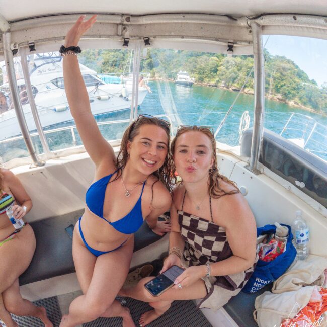 Two smiling women sit on a boat; one in a blue bikini raises her arm and the other in a checkered swimsuit makes a kissy face. The background shows water, boats, and a lush green shoreline.
