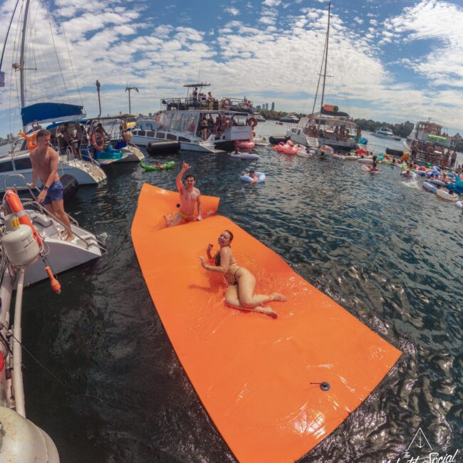 People relaxing on a large orange floating mat in the water, surrounded by boats and other floaties, under a partly cloudy sky. The scene appears festive and social, with several boats anchored nearby.