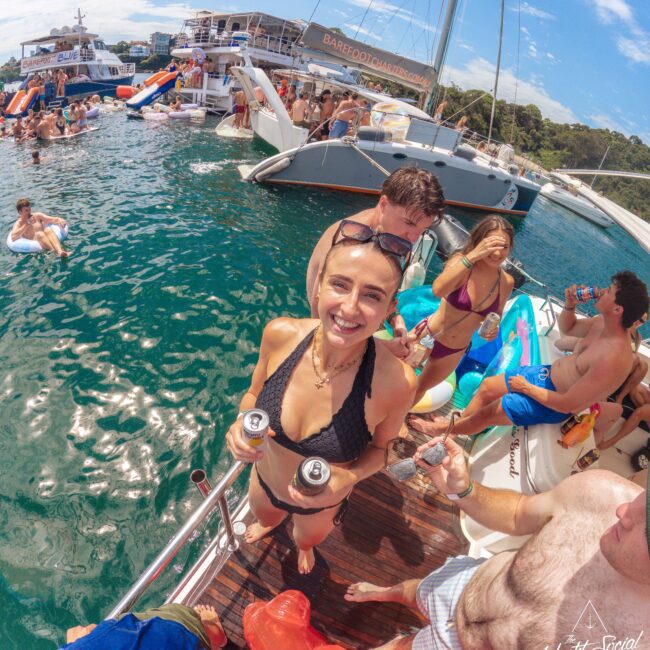 A woman in a black bikini, smiling and holding two cans, stands on a boat with several people socializing around her. Other boats and people swimming or floating on inflatables are visible in the sunny, blue water.