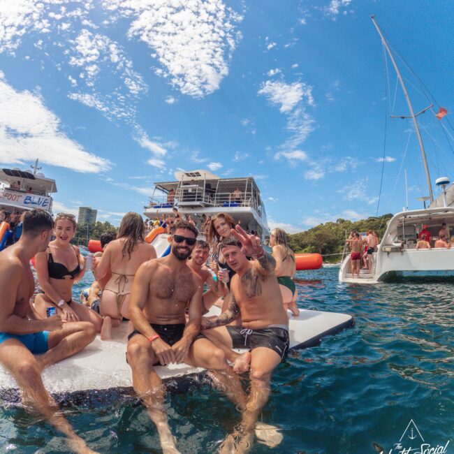 A group of people in swimsuits relax and smile on a floating mat in the water near yachts. Others swim and socialize nearby under a partly cloudy sky, enjoying a lively day on the water.