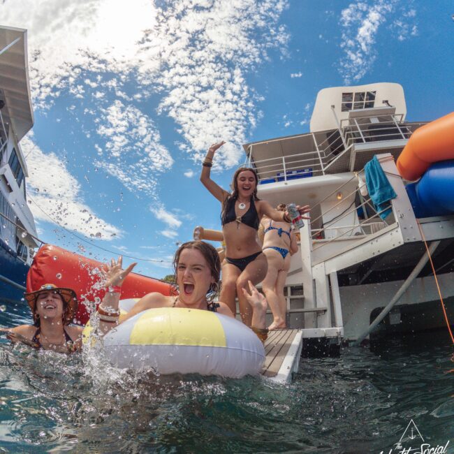 Three women in swimsuits laugh and play on a floating dock by a yacht, with one riding an inflatable pool float in the water. The scene is lively under a bright, partly cloudy sky.