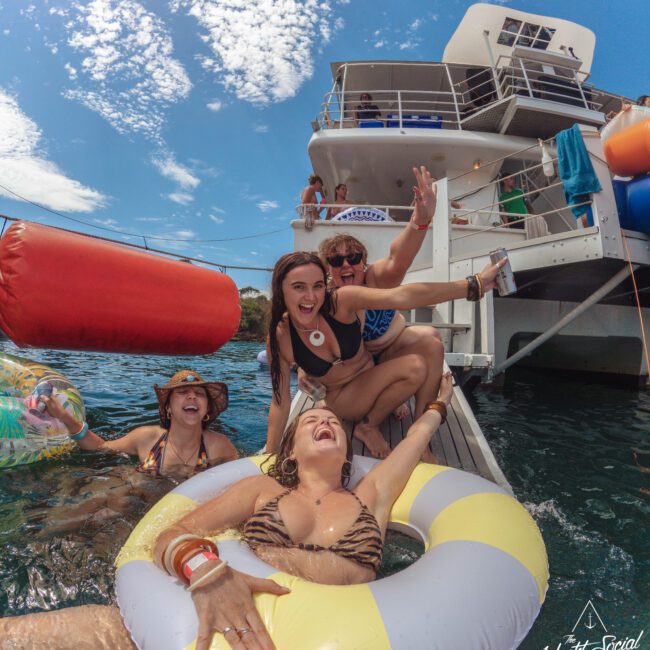 Four women in swimsuits laugh and pose on a dock beside a boat, one leaning back in a yellow-and-white float. The sky is blue with scattered clouds. "Yacht Social Club" is visible in the bottom right corner.
