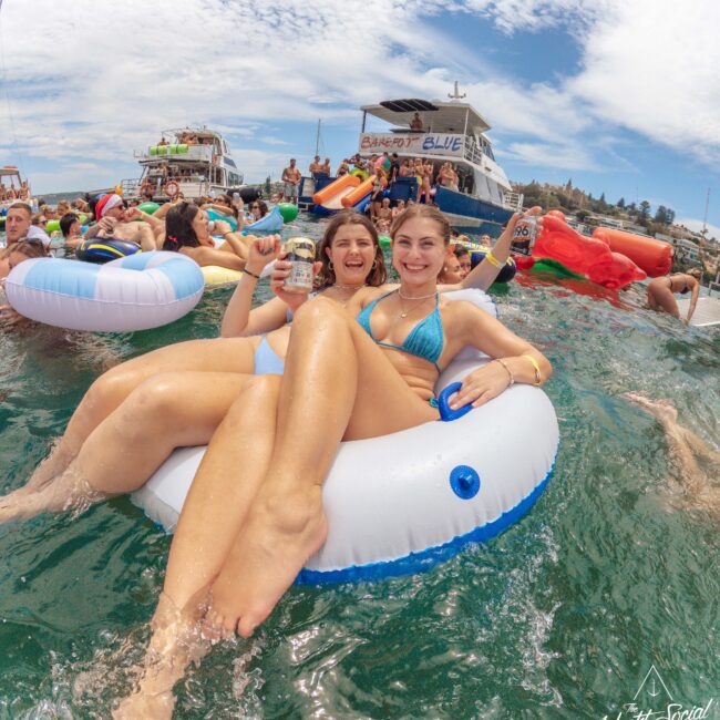 Two women in swimsuits relax on a white inflatable float in the water, smiling and holding drinks, surrounded by other people on floats and boats during a lively summer party.