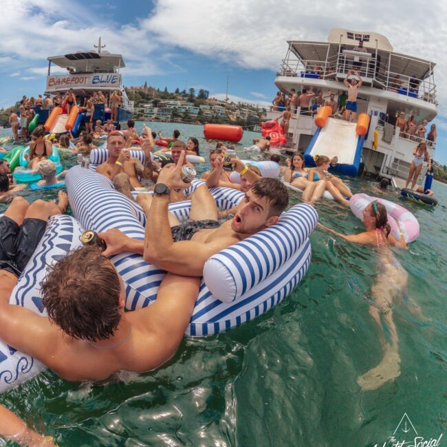 A group of people enjoy a lively party in the water near a boat. Some float on inflatables, one man makes a surprised face, and others swim or relax. The boat in the background features a slide and more partygoers.
