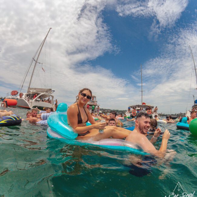 A group of people enjoy a lively boat party, relaxing on colorful inflatables in the water with sailboats anchored nearby, under a partly cloudy sky. Two people smile at the camera on a pool float.