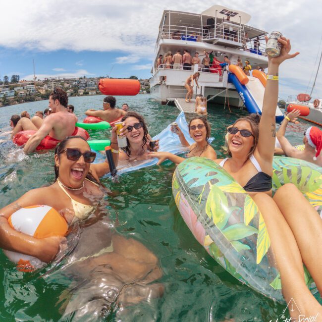 A group of smiling people lounging on colorful floaties and holding drinks in the water near a large party boat, with more people relaxing and playing on the boat under a partly cloudy sky.