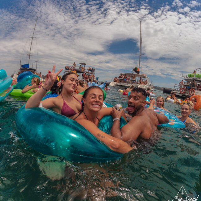 Three people smile and pose for the camera while floating on blue inflatables in the ocean, surrounded by a crowd of others enjoying a lively boat party under a partly cloudy sky. Several boats are visible in the background.
