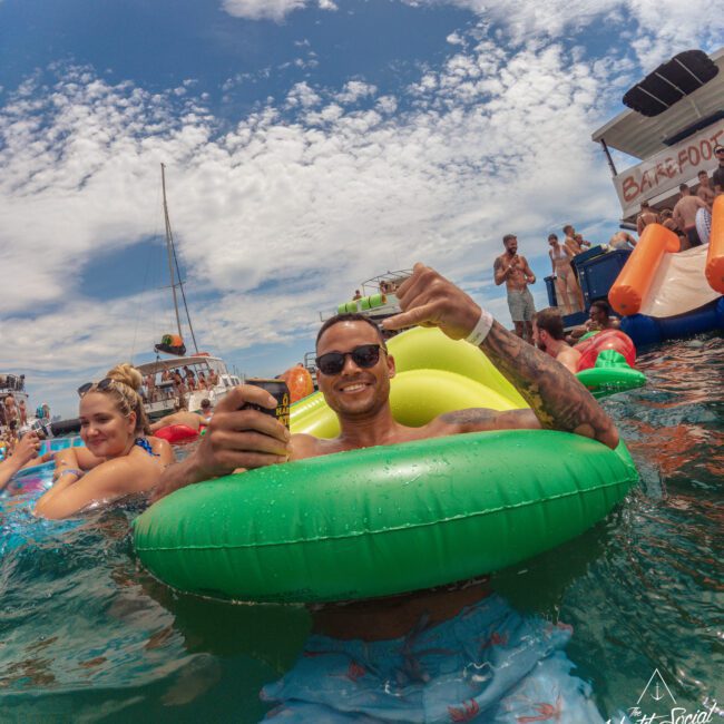 A man floats in a green pool ring, smiling and giving a thumbs-up while holding a drink. Other people relax on colorful inflatables in the water under a partly cloudy sky at a lively outdoor party.