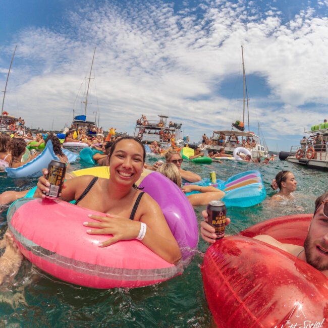 A smiling woman in a pink inflatable ring holds a drink can at a lively boat party in the sea, surrounded by other people on colorful floaties, with boats and a partly cloudy sky in the background.