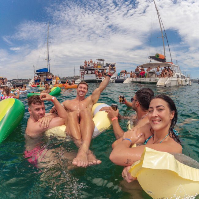 A group of young adults smile and relax on inflatable floats in the ocean, surrounded by others. Boats filled with people are anchored nearby under a partly cloudy sky, creating a festive summer atmosphere.