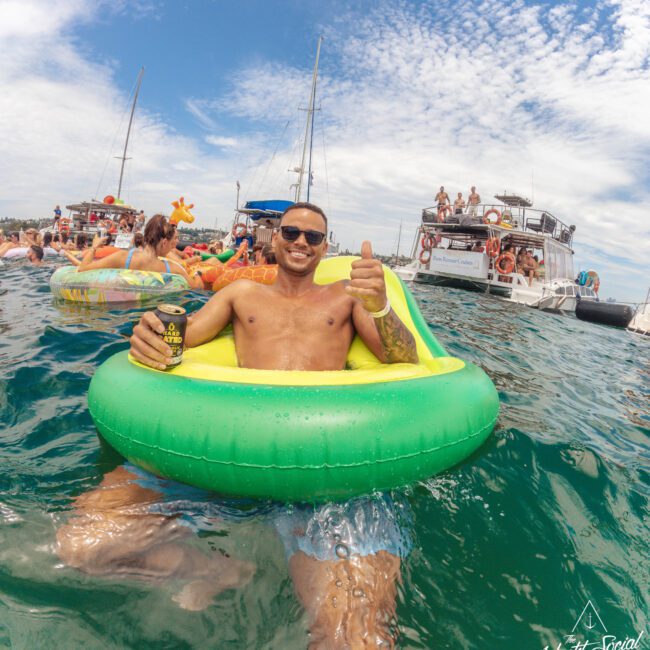 A man wearing sunglasses relaxes in a green inflatable float on the water, holding a drink and giving a thumbs up. Multiple people and boats are in the background under a partly cloudy sky.