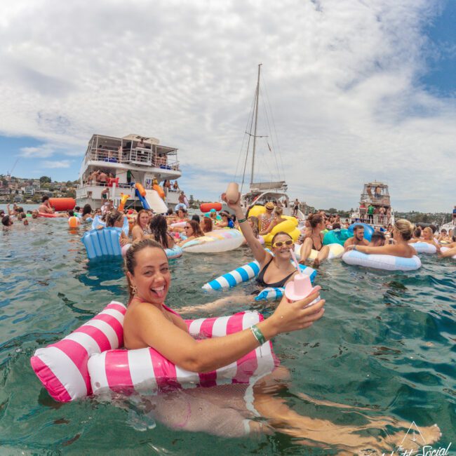 A group of people relax on colorful pool floats in the water, smiling and holding drinks during a lively outdoor party. Boats and a partly cloudy sky are visible in the background.