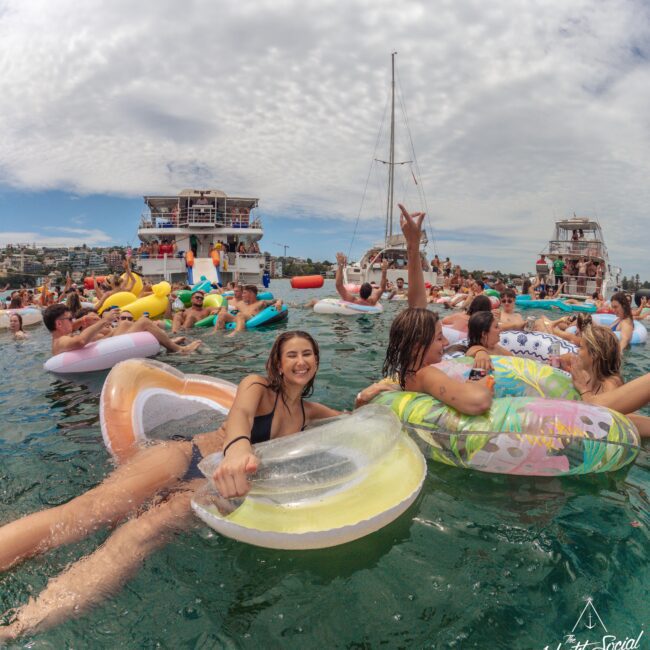 A large group of people float on colorful inflatables in the ocean near yachts, enjoying a lively outdoor party under a partly cloudy sky. Two women in the foreground smile and pose for the camera.