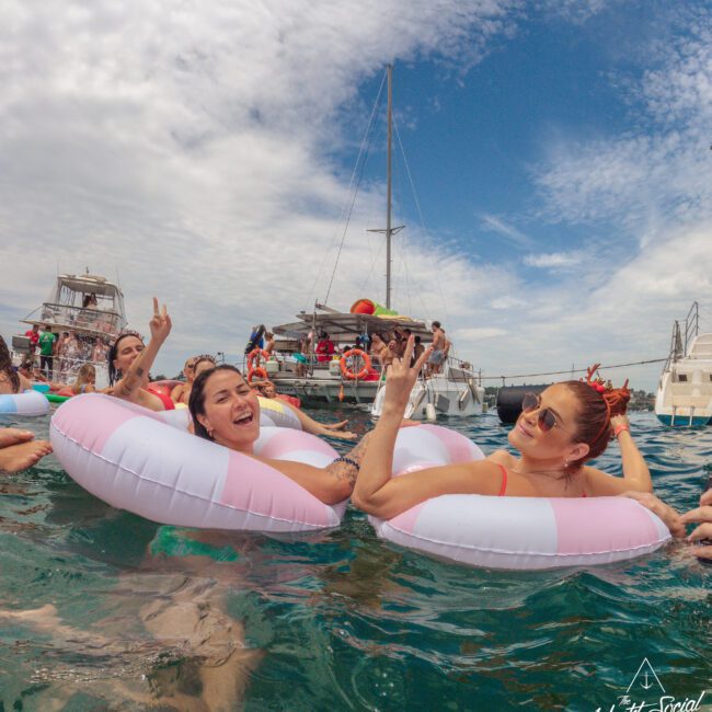 Two women in pink-and-white pool floats smile and gesture to the camera while relaxing in the ocean with other people nearby. Catamarans are anchored in the background under a partly cloudy sky.
