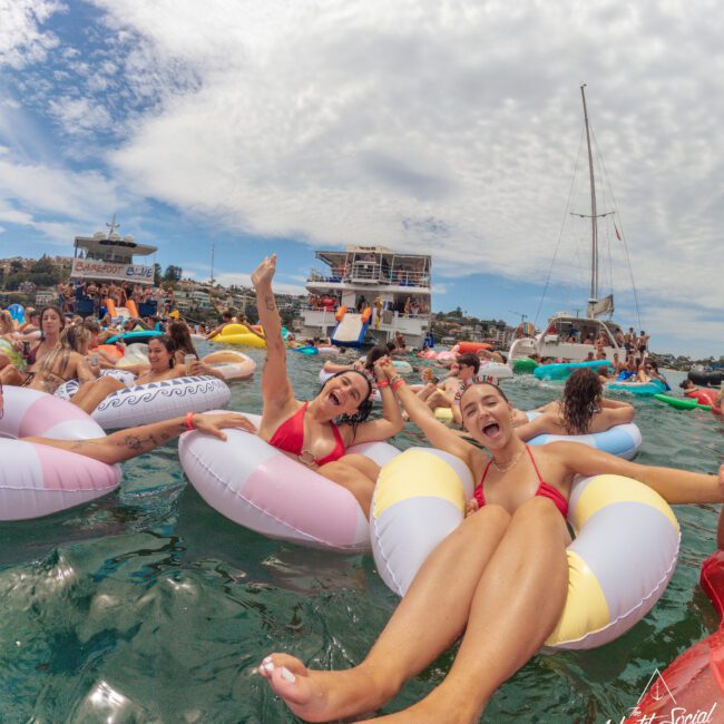 Two women smiling and posing on inflatable pool floats in the water, surrounded by other people on floats. Boats and a yacht are anchored nearby under a partly cloudy sky during a lively summer event.