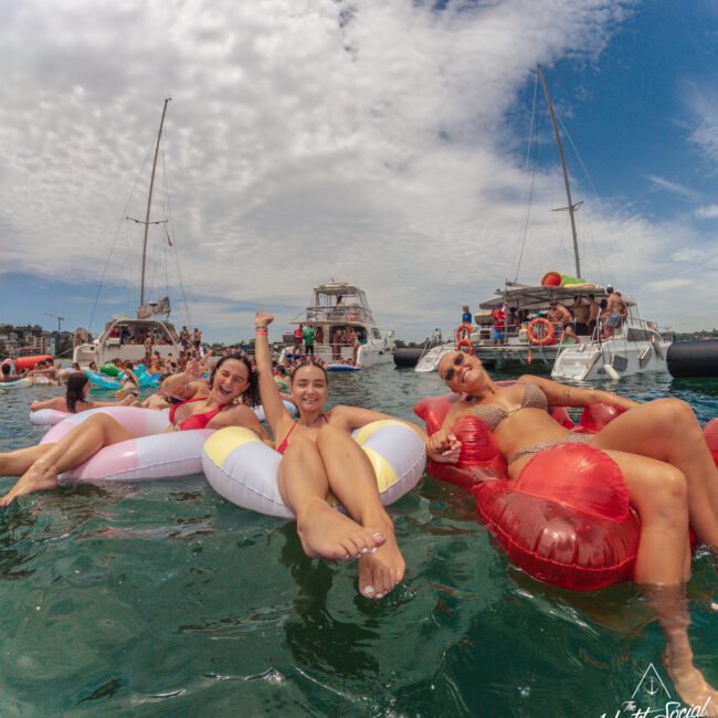 Three women smiling and relaxing on colorful inflatable pool floats in the ocean, with several yachts and groups of people in the background under a partly cloudy sky.